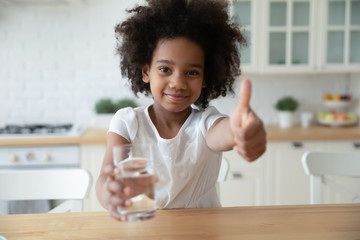Portrait of happy small biracial girl hold water glass show thumb up recommend healthy lifestyle,...