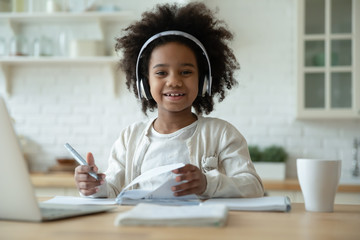 Portrait of smiling little biracial girl in headphones do homework study online in kitchen, happy...