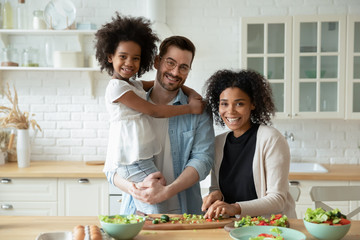 Portrait of overjoyed young multiracial family with little daughter posing in modern kitchen...