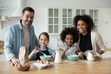 Portrait of happy young multiracial family with little daughters bake biscuits in kitchen together, smiling parents with small multinational daughters children have fun cooking breakfast at home
