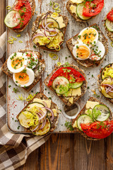 A set of breakfast sandwiches made from homemade buckwheat bread with various toppings on a wooden rustic board, top view.