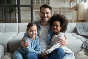 Portrait of young caring father sit on couch hug cuddle with small multiracial daughters, happy Caucasian dad embrace play with multiethnic little children, enjoy family weekend at home together