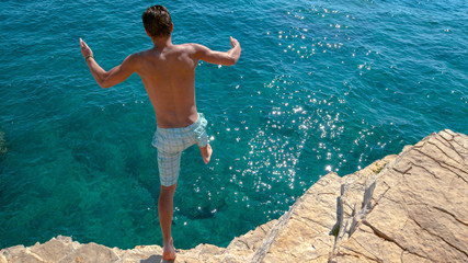 Carefree man dives off a towering rocky cliff and into the deep blue sea.