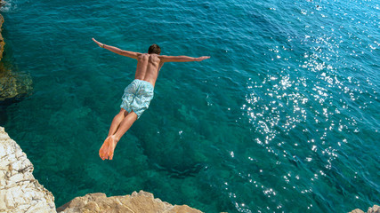 COPY SPACE: Man jumping off a rocky ledge and into the glistening blue ocean.