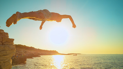 CLOSE UP: Golden sunset shines on rock diver jumping off a cliff and into sea.