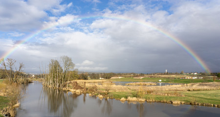 Feld entlang der Mainaue (Fluss) mit Regenbogen vor blauem Himmel leuchtet über dem Panorama. Mainaue. Bayreuth. Deutschland.
