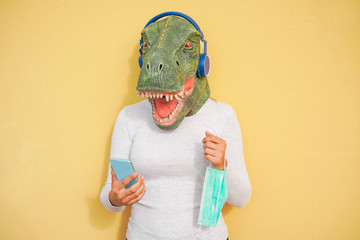 Crazy woman wearing t-rex and holding protection mask while listening music with phone app -...