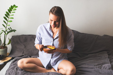 Young woman holding a plate with smothie bowl with fruit and yogurt, sitting on bed, home