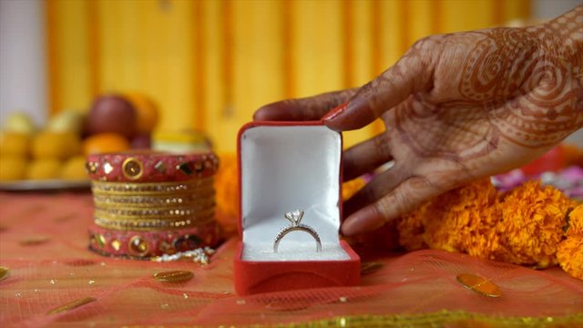 Closeup of mehndi on woman hands placing a red box of her engagement ring. Beautiful Indian bride's wedding ring placed on a decorated platform with bangles  Chooda  mangal sutra  fruits  and sweets
