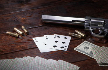 Gambling in cards, poker or blackjack for money. On a wooden table are a pistol with cartridges, cash dollars, and a deck of playing cards with an ACE of spades and a six. Dangerous gambling debts