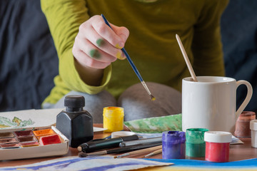 A girl whose face is not visible draws at the table covered with drawing accessories - paper, brushes, paint cans and others.