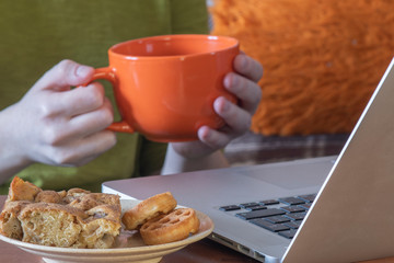 A girl whose face is not visible is sitting at a laptop with a cup of hot drink and cookies. Cozy pastime.