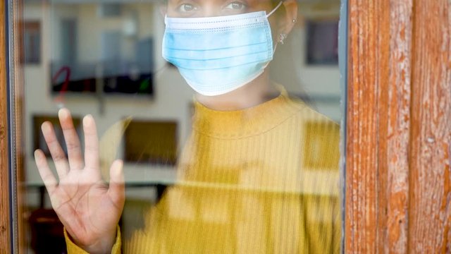 Close up of a hopeful young caucasian south american girl with dark hair yellow shirt ank blue mask on looking outside her window and posing her hand on it during covid-19 lockdown