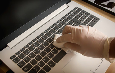 Woman cleaning the laptop at home wearing gloves and using a disinfecting wipe to ensure the...