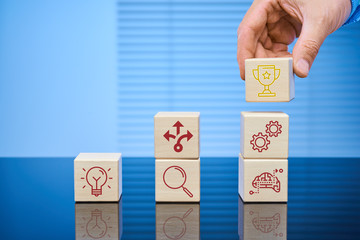 Visual business concept of the process from idea to result. Wooden cubes on a blue background with icons of various actions. The male hand sets a cube with the goal achievement icon on top.