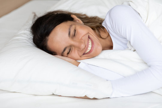 Close up happy woman with healthy toothy smile resting under warm blanket in comfortable bed, enjoying early morning after awakening, beautiful girl with closed eye lying on soft pillow in bedroom