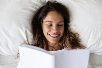 Top view close up smiling young woman reading book, resting in comfortable bed in the morning, happy beautiful girl enjoying favorite novel, literature, relaxing on soft pillow, leisure concept