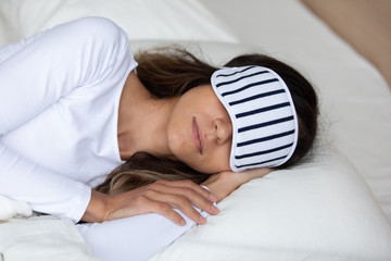 Woman wearing striped sleeping mask and pajama resting on soft pillow with hand under cheek close...