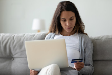 Serious woman holding credit card, reading information, using laptop, sitting on sofa at home,...