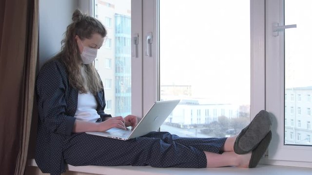 Woman wearing mask and working on laptop from home due to coronavirus self isolation. Sitting on window sill. Authentic home workplace. Handheld shot with gimbal. Coronavirus outbreak 2020.