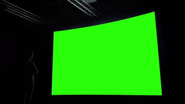Woman looking at large wall blank interactive green display in dark room of modern technology exhibition, cinema or museum. Mock up, green screen, futuristic, template, education, chroma key concept