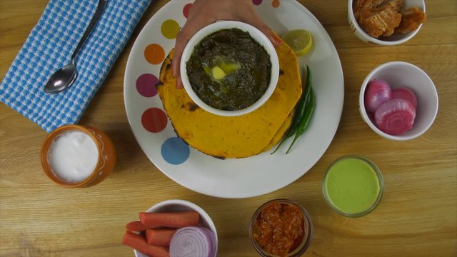 Woman hands placing a bowl of freshly prepared Sarson ka saag in a ceramic plate. Popular Punjabi cuisine Makki ki Roti and saag served with Lassi  salad  pickle  mint chutney  and jaggery - winter...