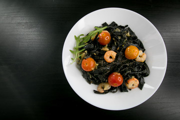 Black Spaghetti or tagliatelle with shrimps, cherry tomato and basil served in a white plate on a dark wooden table. Italian cuisine