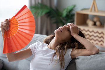Fototapeta Overheated woman sitting on couch, waving orange paper fan close up, girl feeling unwell, suffering from heating at home, feeling discomfort, hot summer weather or fever, sitting on couch alone obraz