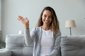 Happy young woman with toothy smile waving hand, looking at camera, sitting on couch at home, vlogger recording webinar, using webcam, chatting online, making video call, remote job interview
