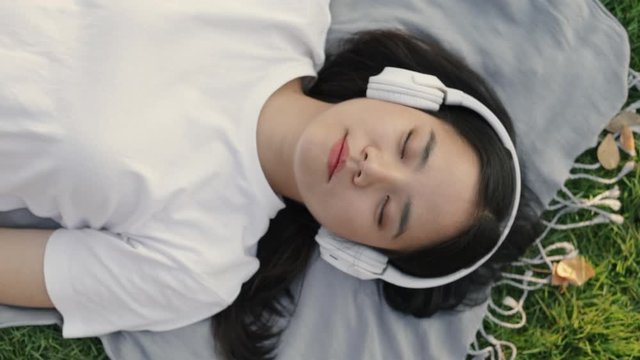 Asian female wearing headphones listening to music while lying on the floor grass outdoors at a public park on the beautiful sunset. Feeling a freedom relaxing lifestyle concept.