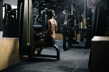 Fototapeta na wymiar Fitness young woman with perfect athletic body wearing black sportswear sitting at simulator and prepares for training in modern dark gym. Concept of healthy lifestyle.
