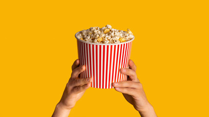 Unrecognizable man holding striped paper bucket with popcorn on orange background, close up....