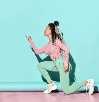 Young beautiful girl with blond dreadlocks hairstyle in sexy sportswear and sneakers sitting on floor and dancing