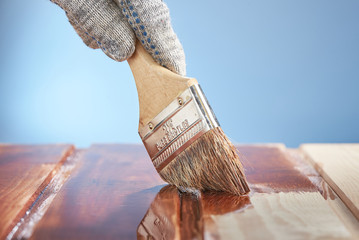 Man's hand with a paint brush painting wooden surface on a blue background.
