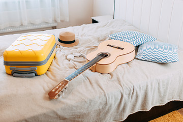 Yellow suitcase, white guitar and hat on the bed, summer vacation during quarantine and coronavirus, holidays while quarantine and self-isolation