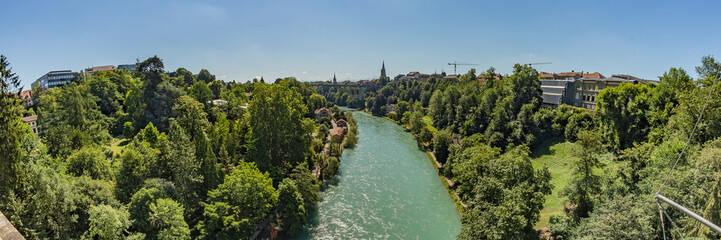 Fototapeta na wymiar Bern, Switzerland - July 30, 2019: Panoramic view of Aare river from the Lorrainebrucke Bridge at sunny summer day. Snow-capped peaks of the Alps in a light haze in the background