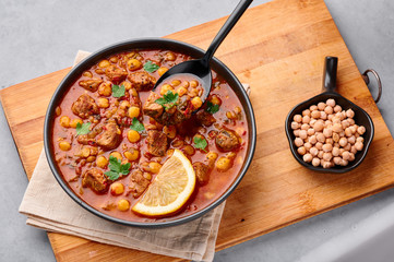 Moroccan Harira Soup in black bowl at grey concrete table top. Harira is Moroccan Cuisine dish with lamb or beef meat, chickpeas, lentils, tomatoes, ciliantro. Ramadan Iftar Food.