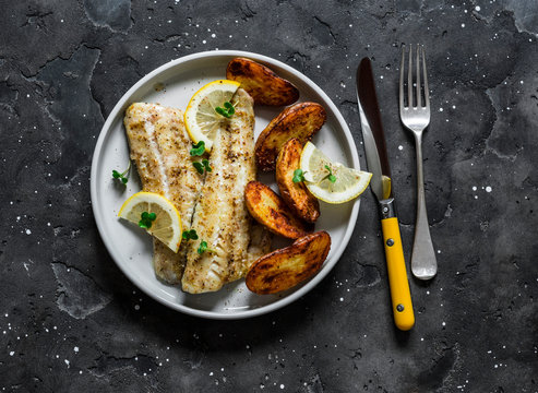 Fish fillet baked with lemon and new potatoes on a dark background, top view. Delicious homemade lunch