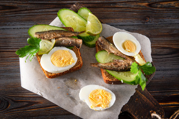 rye bread sandwiches with sprats, fresh cucumber, herbs and boiled egg on a dark wooden background, canned fish,