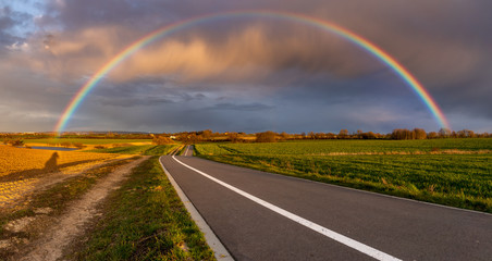 colorful rainbow against the backdrop of dramatic, storm clouds over a spring field and bicycle path