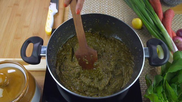 Pan shot of hands cooking Sarson ka saag on induction cooker - Punjabi cuisine. Closeup shot of an Indian female stirring traditional Punjabi food saag in a pan surrounded with vegetables on a kitc...