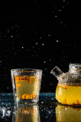 herbal sea buckthorn tea in a transparent teapot and in a transparent glass with a double bottom on a black background with splashes of water
