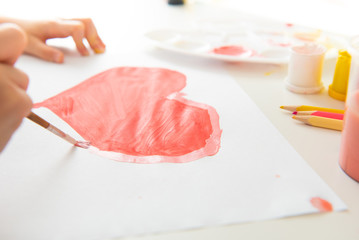 child draws a pink heart in gouache on a white table with a palette