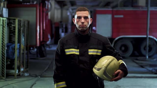Firefighter video portrait wearing uniform and holding yellow protective helmet. Fire trucks in the background. Camera moving left.