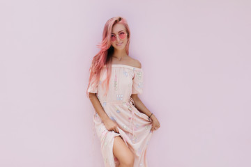 Carefree girl in stylish summer dress posing with interest smile. Inspired caucasian woman with pink hair standing on white background.