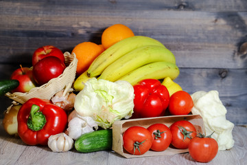 On a gray graphite wooden background, tomatoes in a box, cucumber, garlic, iceberg lettuce, large red peppers, bananas, oranges in a wicker container red apples.