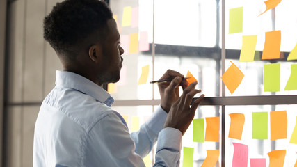Focused African American male employee write on colorful sticky notes develop business project in...