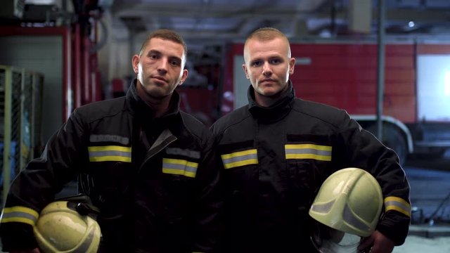 Two firefighters standing together, wearing uniform and holding yellow protective helmets. Fire trucks in the background. Camera moving left.