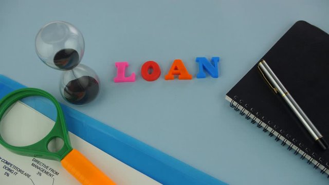 Closeup shot of the word "Loan" composed with colorful letters - microfinance concept. Pan shot of plastic letters spelling the word "Loan" surrounded by a sand clock  papers  file  diary  pen  and...