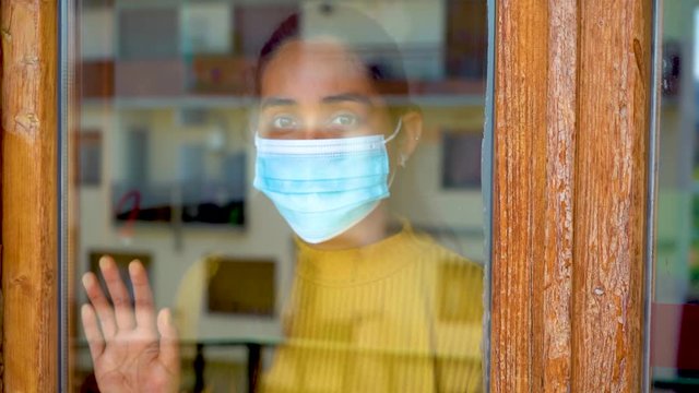 Hopeful eyes of young caucasian south american girl with dark hair yellow shirt ank blue mask on looking outside her window and posing her hand on it during covid-19 lockdown